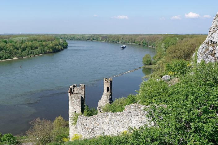 A broken stone curtain wall and a small ruined tower standing on a rocky spur high above a wide river, dense green woodland on both banks and a boat moving upstream in the distance.