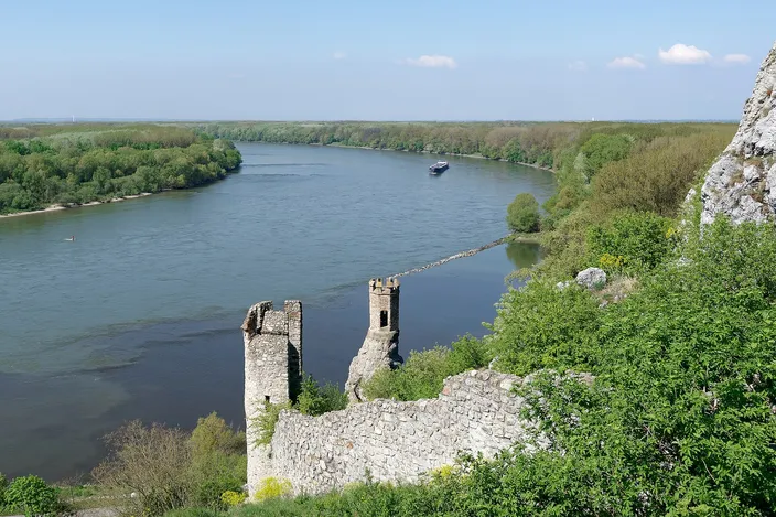 A broken stone curtain wall and a small ruined tower standing on a rocky spur high above a wide river, dense green woodland on both banks and a boat moving upstream in the distance.