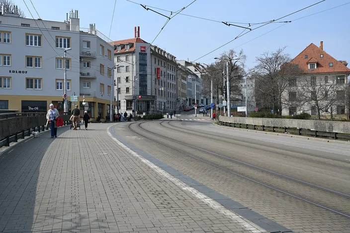 Zámocká Street curving away below the castle hill in Staré Mesto: pale early-20th-century apartment blocks, tram tracks and overhead wires, a cobbled pedestrian walkway with a few people strolling, bare trees on a clear day.