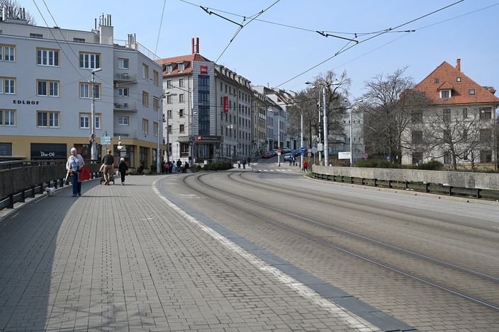 Zámocká Street curving away below the castle hill in Staré Mesto: pale early-20th-century apartment blocks, tram tracks and overhead wires, a cobbled pedestrian walkway with a few people strolling, bare trees on a clear day.