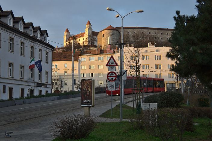 A red Bratislava tram on Kapucínska street in low golden light, with the white boxy castle rising on its hill behind the pale buildings.
