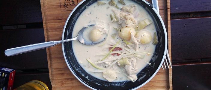 A top-down bowl of pale, creamy chicken waterzooi with pieces of leek, potato and chicken and a spoon resting in it