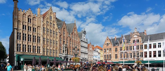 Bruges Markt square with stepped-gable brick houses, a horse-drawn carriage and café terraces under a blue sky with clouds