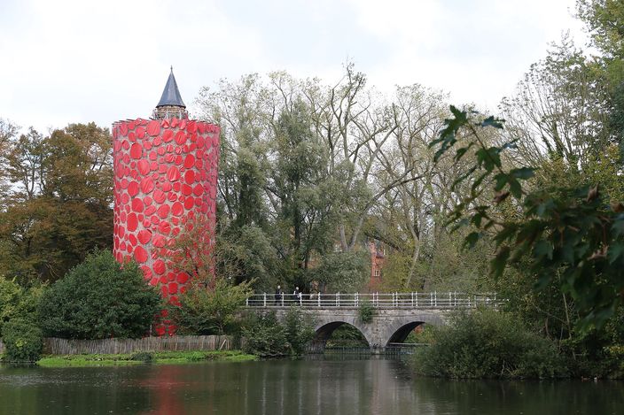 A still canal basin at the Minnewater in Bruges, with a low stone arched bridge, willows and trees along the banks, and a round medieval brick tower on the left wrapped in a temporary red artwork of overlapping discs beneath its slate spire.