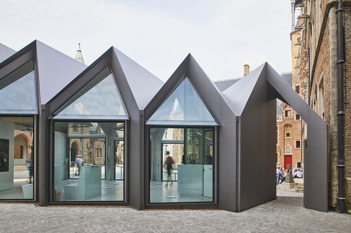 The Gruuthusemuseum's entrance pavilion in Bruges: a low modern building of dark metal with steep glazed gables standing on cobbles, the medieval brick palace with its Gothic windows and red door beside it.
