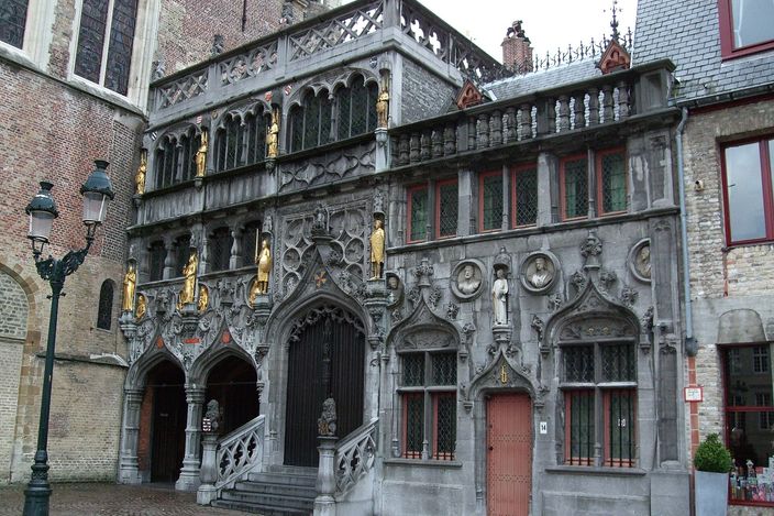 The ornate grey stone facade of the Basilica of the Holy Blood on the Burg in Bruges, with gilded statues standing in canopied niches, a covered outer staircase and Gothic arched doorways above cobbles.