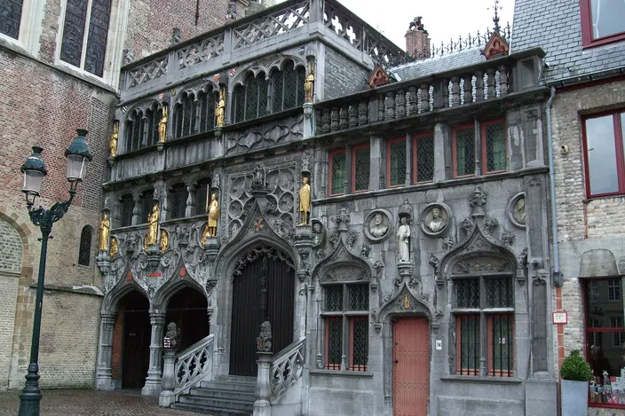 The ornate gray stone facade of the Basilica of the Holy Blood on the Burg in Bruges, with gilded statues standing in canopied niches, a covered outer staircase and Gothic arched doorways above cobbles.