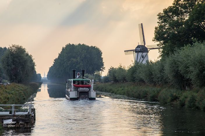 A straight tree-lined canal in hazy early-morning light, with a small white paddle steamer coming up the water and the whitewashed tower of the Schellemolen windmill standing among poplars on the right bank.