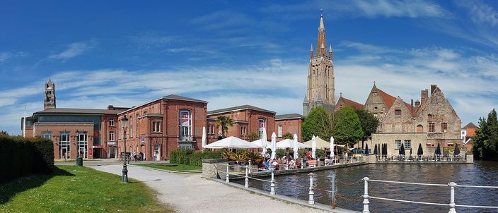 A canal in Bruges with a long red-brick building and a row of white parasols along its bank, a gravel path and iron railings in the foreground, and the tall brick tower and spire of the Onze-Lieve-Vrouwekerk rising behind gabled houses under a blue sky.