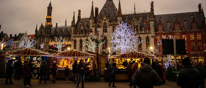 Lit wooden market huts on the Markt in Bruges at dusk, white light-trees between them and the neo-Gothic Provincial Court rising behind
