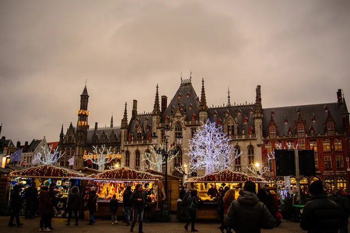 Lit wooden market huts on the Markt in Bruges at dusk, white light-trees between them and the neo-Gothic Provincial Court rising behind