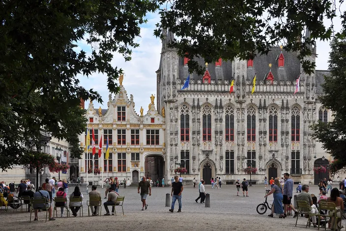 The Burg square in Bruges in daylight, with the ornate Gothic City Hall and the flag-hung Civil Registry facade behind a leafy tree, people seated and strolling across the cobbles.