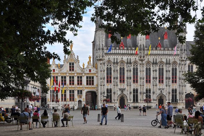 The Burg square in Bruges in daylight, with the ornate Gothic City Hall and the flag-hung Civil Registry facade behind a leafy tree, people seated and strolling across the cobbles.