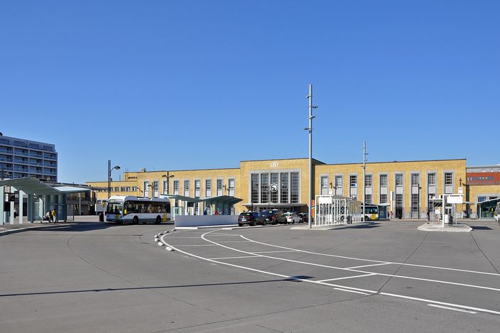 The yellow-brick facade of Bruges railway station across its bus forecourt, with a De Lijn city bus at the stops under a clear blue sky.