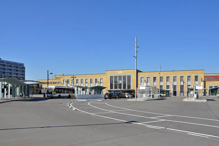 The yellow-brick facade of Bruges railway station across its bus forecourt, with a De Lijn city bus at the stops under a clear blue sky.