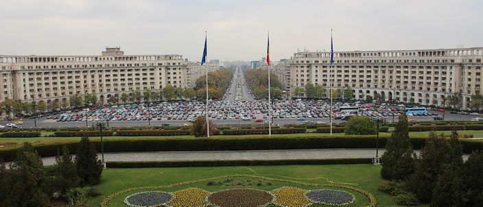 Elevated view from the Palace of the Parliament looking east: a vast paved square filled with parked cars, three flagpoles, and two long symmetrical government blocks flanking a straight boulevard that runs to a hazy horizon, with a circular flowerbed on the lawn in the foreground