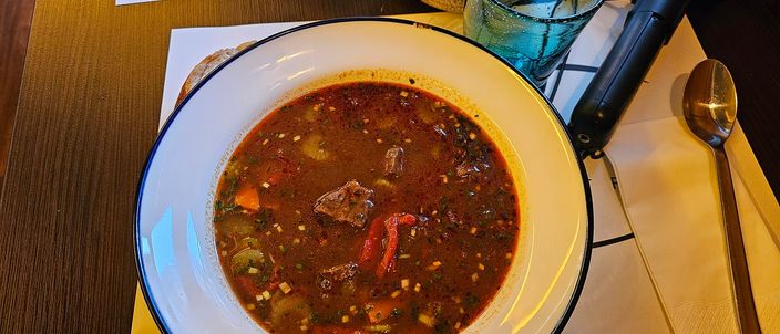 A bowl of gulyás soup: a red paprika broth with chunks of beef, potato and vegetables, served with cutlery on a table.