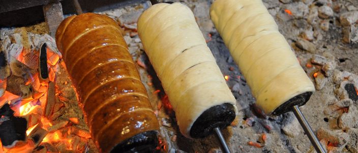 Two kürtőskalács baking on spits over glowing charcoal, their sugar coating turned golden brown.