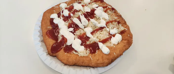 A golden, deep-fried lángos on a paper plate, puffed and blistered, topped with soured cream and grated cheese.
