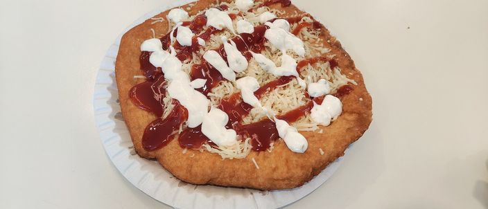 A golden, deep-fried lángos on a paper plate, puffed and blistered, topped with soured cream and grated cheese.