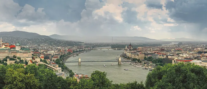 Panorama from Gellért Hill over the Danube: Buda Castle on the left bank, the Chain Bridge spanning to Pest, Parliament's dome on the right, hills and a dramatic sky behind.