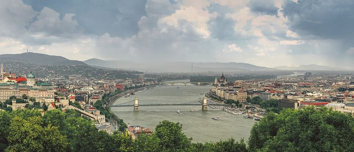 Panorama from Gellért Hill over the Danube: Buda Castle on the left bank, the Chain Bridge spanning to Pest, Parliament's dome on the right, hills and a dramatic sky behind.