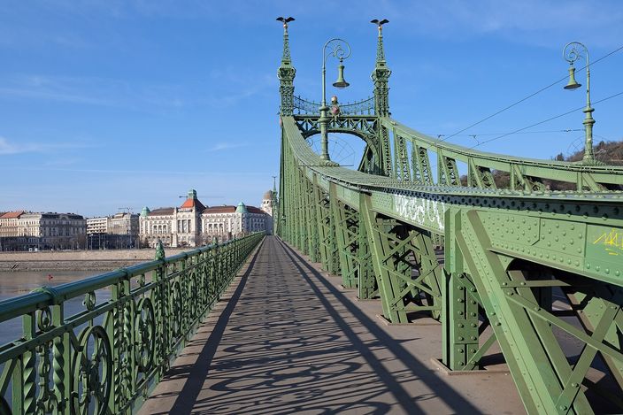 The pedestrian walkway of Budapest's Liberty Bridge, running away from the camera between pale green riveted ironwork trusses and lamp-topped masts, with the Danube and the Pest embankment buildings to the left under a clear blue sky.