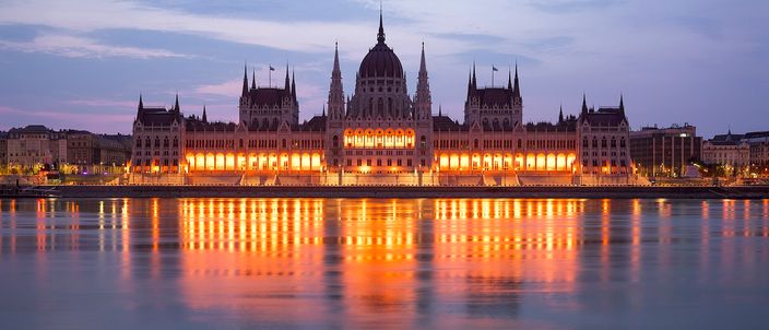 The Hungarian Parliament Building seen from the far bank of the Danube at dusk, its long neo-gothic river facade and red dome floodlit gold, with the lit windows reflected in a band down the still water.
