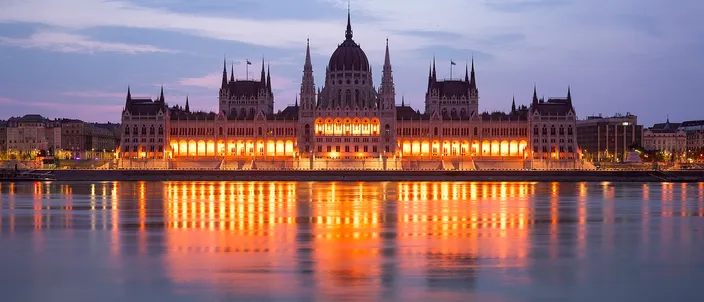 The Hungarian Parliament Building seen from the far bank of the Danube at dusk, its long neo-gothic river facade and red dome floodlit gold, with the lit windows reflected in a band down the still water.