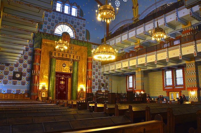 The interior of a synagogue in Hungarian Art Nouveau: a gilded ark under a horseshoe arch flanked by green marble columns, walls of blue and white patterned tiling, brass chandeliers hanging from a starred vault, and dark wooden pews in rows.