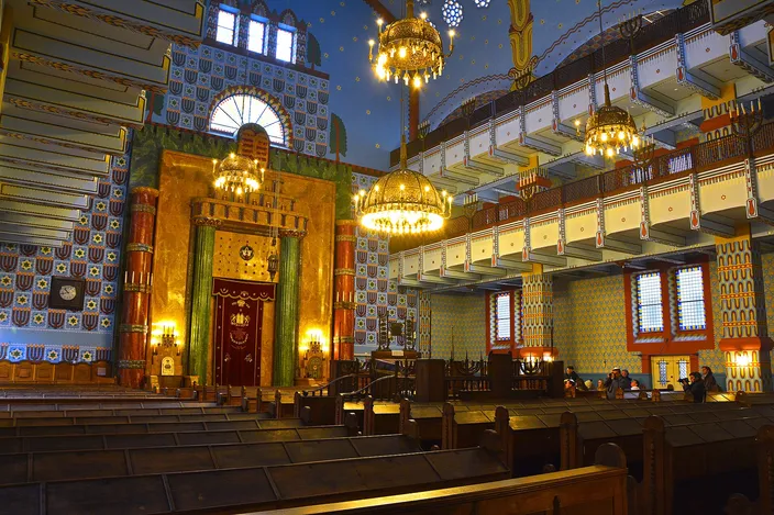 The interior of a synagogue in Hungarian Art Nouveau: a gilded ark under a horseshoe arch flanked by green marble columns, walls of blue and white patterned tiling, brass chandeliers hanging from a starred vault, and dark wooden pews in rows.