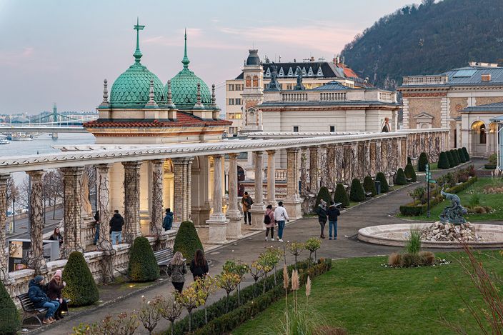 The terraces of the Varkert Bazar at dusk: a long colonnade of pale stone columns running downhill past two green copper domes, clipped shrubs and a fountain, a few people walking the paths, with the Danube and a bridge beyond on the left and a wooded hillside on the right.
