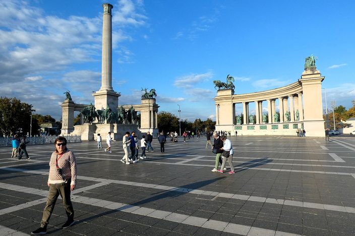 Heroes' Square in Budapest: a tall stone column topped by a winged figure, mounted bronze chieftains at its base, and a curved colonnade of statues to the right, seen across the wide patterned paving with people crossing it under a blue sky with white cloud.