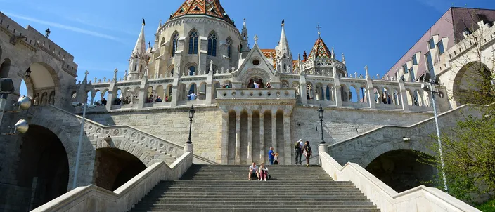 The white stone turrets and arcaded terraces of the Fisherman's Bastion rising above a broad flight of steps, with the patterned tiled roof and pale spires of Matthias Church behind them against a clear blue sky.