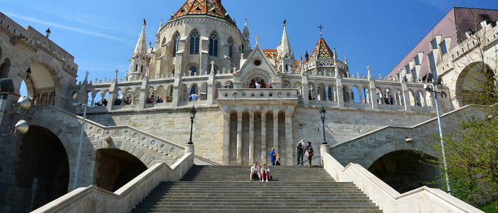 The white stone turrets and arcaded terraces of the Fisherman's Bastion rising above a broad flight of steps, with the patterned tiled roof and pale spires of Matthias Church behind them against a clear blue sky.