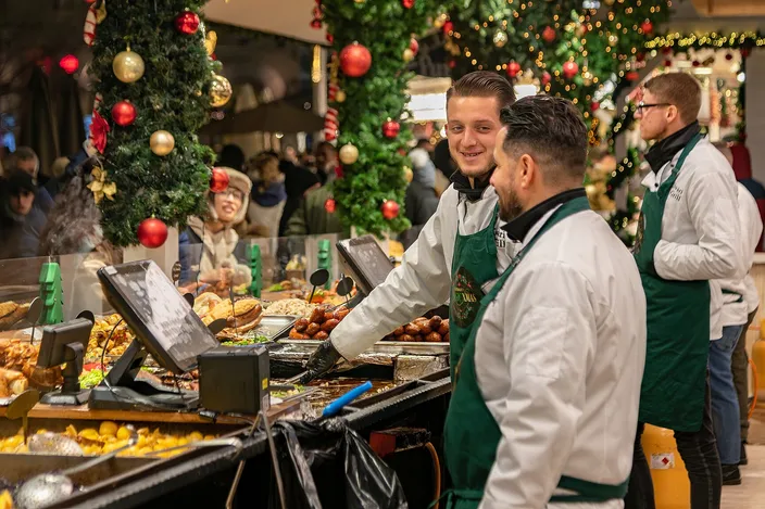 Three cooks in green aprons serving at a market kitchen counter piled with roast meats and potatoes, a fir garland with baubles hanging over the stall