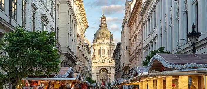 Market stalls lining a street of grand apartment fronts in Budapest's inner city, a chimney-cake stall in the foreground and the dome and front of St Stephen's Basilica closing the view