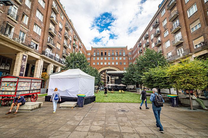 A paved square in Budapest's Jewish Quarter framed by red-brick apartment blocks with an archway through the centre; a market bookstall on a wooden cart to the left, a white pop-up tent, patch of grass and a few pedestrians crossing under a blue sky.