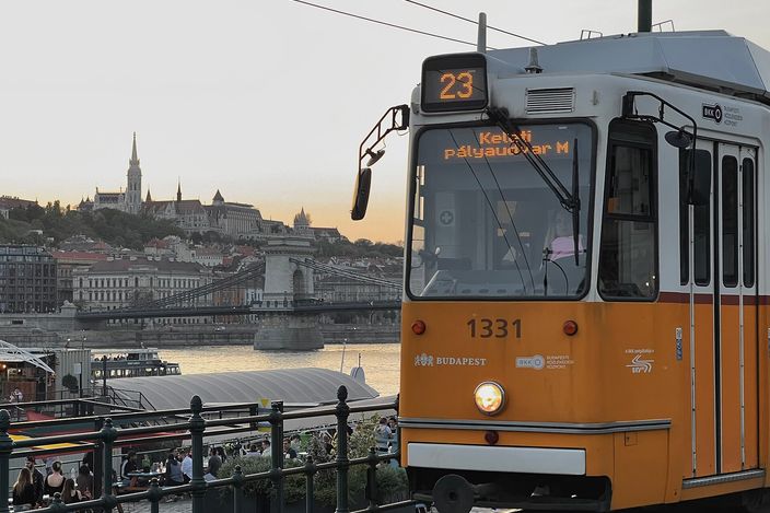 A yellow Budapest tram (line 23, number 1331) crossing along the Pest embankment at dusk, with the Chain Bridge, the Danube and Buda Castle hill with Matthias Church glowing behind it.