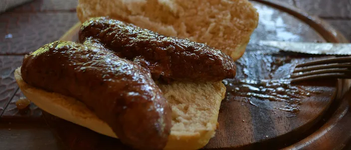 An Argentine choripán on a wooden board: a butterflied grilled chorizo, browned and charred from the grill, laid in a split crusty bread roll