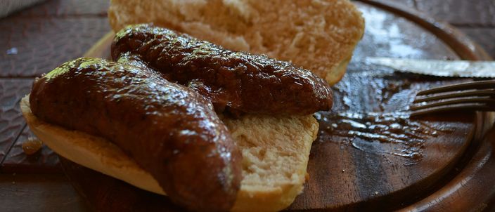 An Argentine choripán on a wooden board: a butterflied grilled chorizo, browned and charred from the grill, laid in a split crusty bread roll