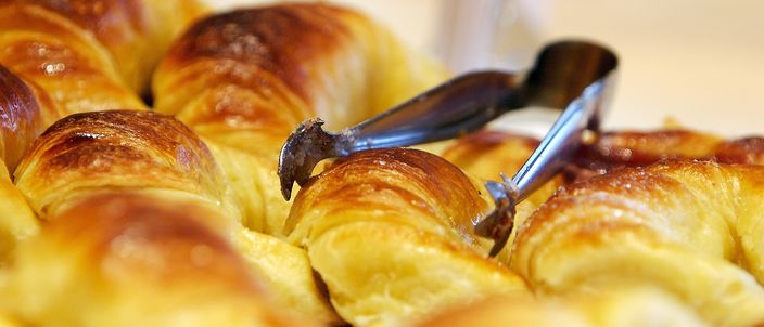 A tray of freshly baked Argentine medialunas, small glossy golden crescents with a sugar-syrup glaze, served with tongs