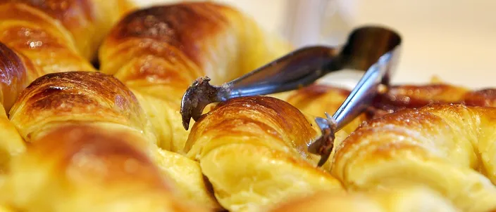 A tray of freshly baked Argentine medialunas, small glossy golden crescents with a sugar-syrup glaze, served with tongs