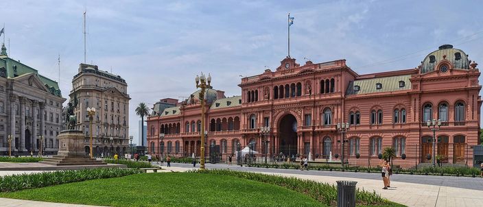 Panorama of Plaza de Mayo with the pink Casa Rosada on the right, the domed Banco de la Nación and a monument column, under a bright sky with people crossing the plaza