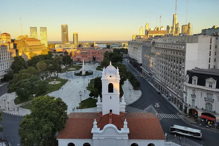 Late-afternoon aerial over Plaza de Mayo: the white colonial Cabildo with its bell tower and tiled roof in the foreground, the plaza's paths, palms and central monument behind it, the pink Casa Rosada closing the far end, glass towers catching low gold sun on both sides and red buses on the avenue to the right.
