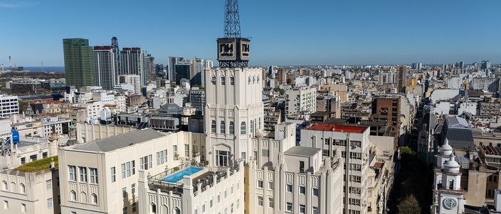 Aerial view over the dense flat rooftops of the Buenos Aires historic centre, a white stepped art-deco tower with a dark metal lattice crown standing above them, a straight street cutting away to the right past a green church dome, and the low city running to a hazy horizon under a clear blue sky.