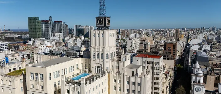 Aerial view over the dense flat rooftops of the Buenos Aires historic center, a white stepped art-deco tower with a dark metal lattice crown standing above them, a straight street cutting away to the right past a green church dome, and the low city running to a hazy horizon under a clear blue sky.