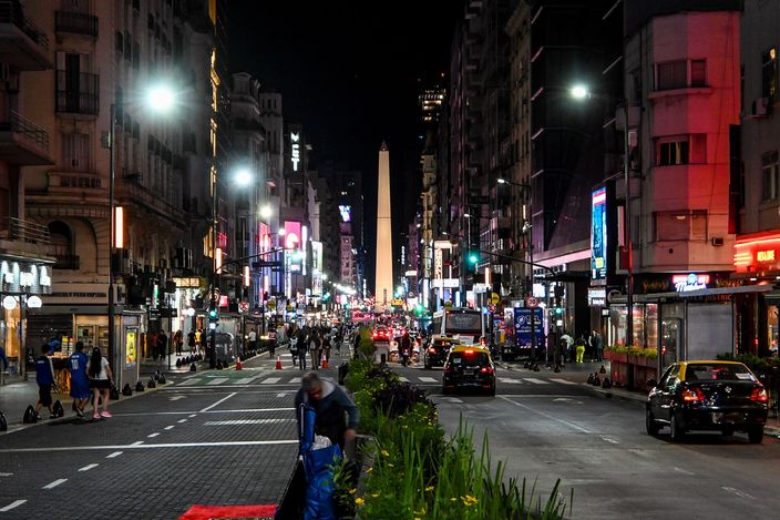 Avenida Corrientes at night, looking down the wide street between lit shopfronts and theatre frontages, cars and pedestrians on the roadway, and the floodlit white obelisk standing at the far end between the buildings.
