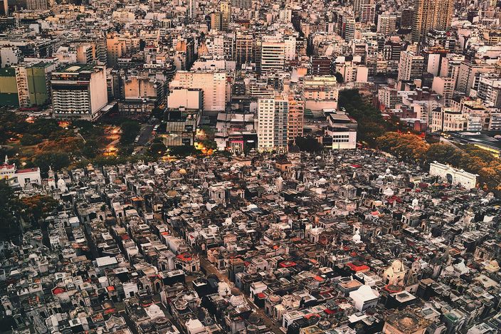 Aerial view over the Recoleta cemetery at dusk: hundreds of stone mausoleums packed along a grid of narrow lanes, one of them carrying a gilded dome, with the dense blocks and towers of Buenos Aires rising immediately beyond the boundary trees under an orange and grey sky.