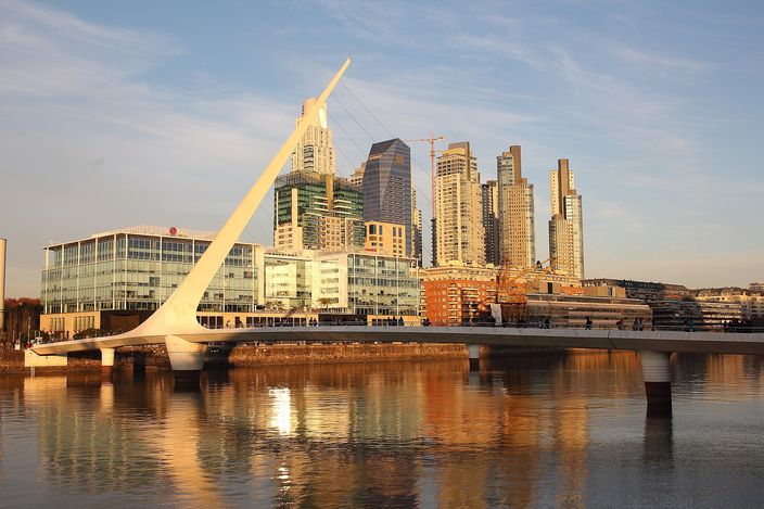 The white Puente de la Mujer footbridge across a Puerto Madero dock at golden hour, its single mast leaning steeply out over the water with cables fanning down to the deck, glass towers and a dockside crane behind it, the whole span mirrored in still water.